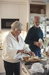 Ältere Frau bereitet Essen zu, während ihr männlicher Partner in der Küche ein Getränk hält - MASF20003