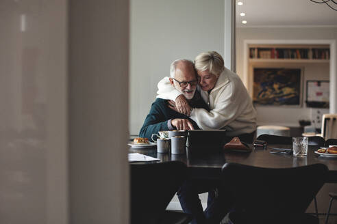 Lächelnde ältere Frau, die ihren Partner am Esstisch im Wohnzimmer umarmt - MASF20004