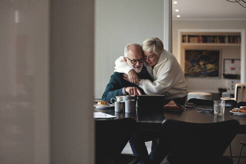 Smiling senior woman embracing partner by dining table in living room - MASF20004