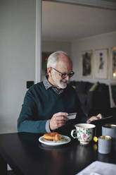 Lächelnder faltiger Mann, der eine Kreditkarte hält, während er ein digitales Tablet auf einem Tisch im Wohnzimmer benutzt - MASF20006