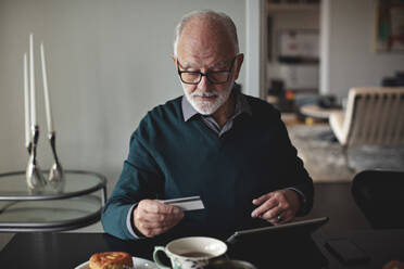 Faltenreicher Mann, der eine Kreditkarte an einem digitalen Tablet hält, während er am Esstisch im Wohnzimmer sitzt - MASF20007