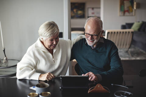 Smiling senior couple using digital tablet while sitting by dining table in living room stock photo