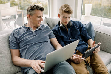 Lächelnder Vater mit Laptop und Blick auf seinen Sohn, der ein digitales Tablet benutzt, während er zu Hause auf dem Sofa sitzt - MASF20031