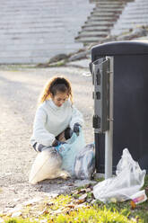 Weiblicher Freiwilliger hockt mit Plastikmüll neben der Mülltonne - MASF20047