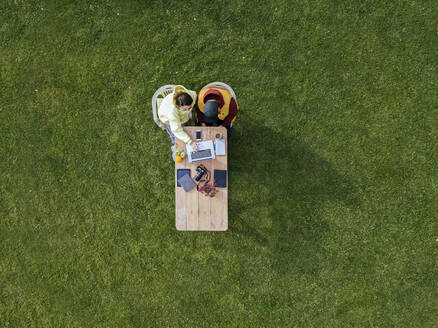 Aerial view of man and woman working together on laptop at coffee table set on green lawn - KNTF05810