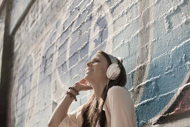 Happy young woman listening music through headphones while standing against wall with graffiti on sunny day - AJOF00300