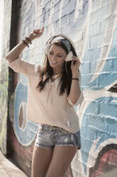 Schöne Frau tanzt beim Hören von Musik über Kopfhörer gegen eine Wand mit Graffiti - AJOF00301
