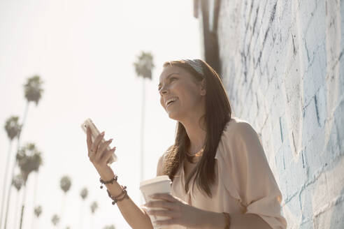 Smiling young woman looking away while holding smart phone and coffee cup - AJOF00302