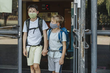 Brothers wearing protective face mask holding hands coming out of school building - MFF06358