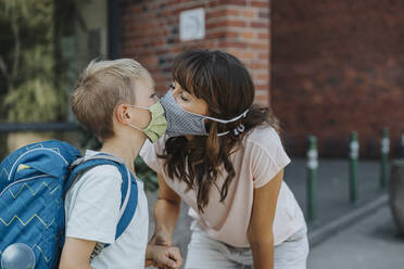 Mother kissing son wearing protective face mask standing in font of school - MFF06360