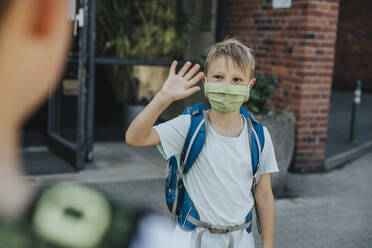 Little boy wearing protective face mask waving his brother outside school building - MFF06361