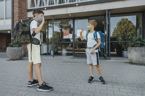 Brothers waving while wearing protective face mask standing in front of school building stock photo