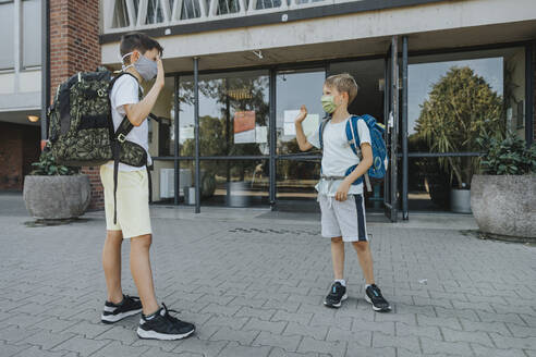 Brothers waving while wearing protective face mask standing in front of school building - MFF06362