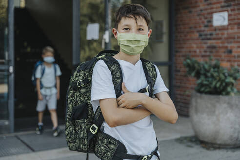 Little boy wearing protective face mask with arms crossed standing in front of school building - MFF06364