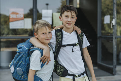 Smiling brothers with arm around standing in front of school building - MFF06365