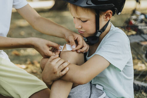 Boy putting bandage on younger brother knee sitting in public park - MFF06376