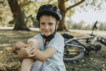 Junge sitzend mit Verband am Knie in einem öffentlichen Park an einem sonnigen Tag - MFF06379