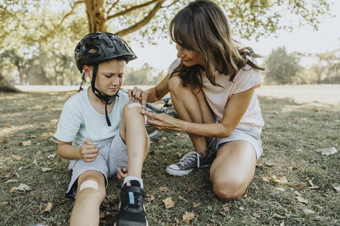 Mother putting bandage on son's knee while sitting in public park during sunny day - MFF06380