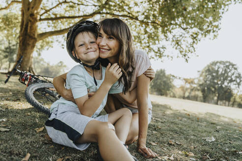 Mother embracing son sitting in public park on sunny day - MFF06381