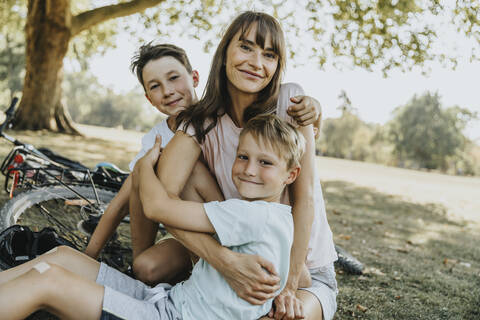 Mutter umarmt ihre Söhne, während sie in einem öffentlichen Park sitzen, lizenzfreies Stockfoto