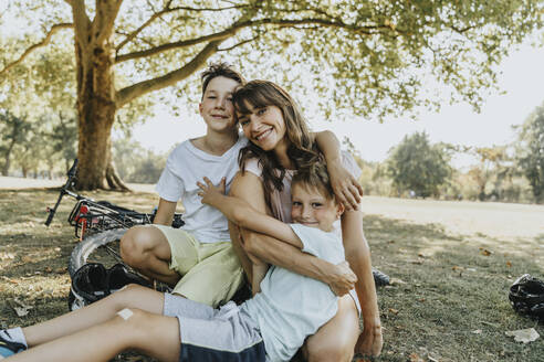 Mother embracing sons while sitting in public park on sunny day - MFF06383