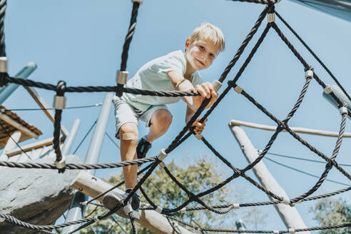 Boy climbing on spider web in public park during sunny day - MFF06388