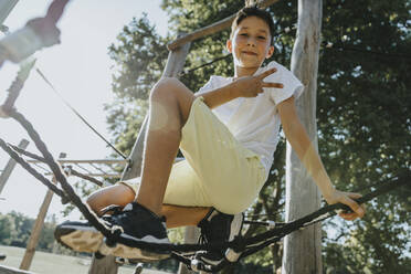 Boy doing hand gesture while sitting on spider web in public park - MFF06389