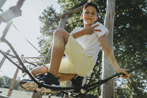 Junge, der eine Handbewegung macht, während er auf einem Spinnennetz in einem öffentlichen Park sitzt - MFF06389