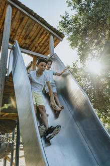 Brothers sliding on slide in public park on sunny day - MFF06392