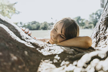 Smiling mature woman with head on tree trunk in public park - MFF06399