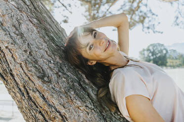 Smiling mature woman with hands in hair lying on tree trunk during sunny day - MFF06402