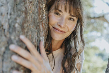 Smiling mature woman leaning on pine tree in public park - MFF06405
