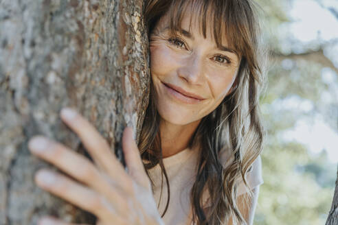 Smiling mature woman leaning on pine tree in public park - MFF06405