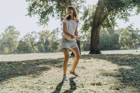 Mature woman throwing frisbee ring while standing in public park on sunny day - MFF06409