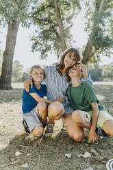 Happy mother embracing sons while kneeling in public park on sunny day - MFF06416