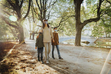 Mutter und Söhne tragen einen Gesichtsschutz beim Spaziergang in einem öffentlichen Park an einem sonnigen Tag - MFF06435