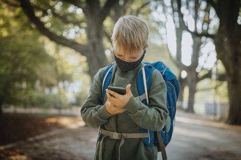 Boy text messaging on smart phone wearing protective face mask while standing in public park - MFF06438