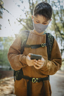 Boy using smart phone wearing protective face mask while standing in public park - MFF06439