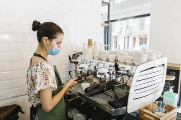 Junge Frau mit Gesichtsmaske beim Kaffeekochen in der Küche eines Cafés - XLGF00595