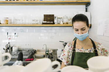 Frau mit Gesichtsmaske bei der Arbeit in der Küche eines Cafés - XLGF00596