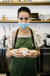 Junge Frau mit Gesichtsmaske und Schürze mit Kaffeetasse in einem Café stehend - XLGF00599