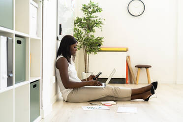 Businesswoman using laptop and mobile phone together while sitting on floor at office - GIOF09272