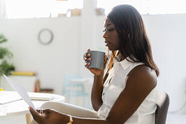 Geschäftsfrau mit Kaffeetasse, die auf einem Stuhl im Büro sitzt und Zeitung liest - GIOF09281