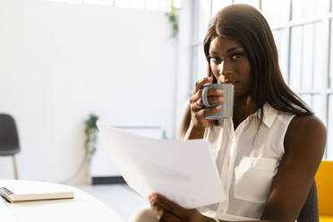 Junge Frau mit Kaffeetasse, die auf einem Stuhl im Büro sitzt und Zeitung liest - GIOF09284