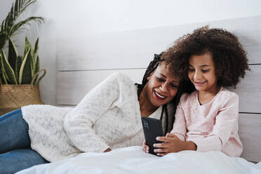 Lächelnde Mutter und Tochter mit Smartphone auf dem Bett im Schlafzimmer zu Hause sitzend - EBBF00930