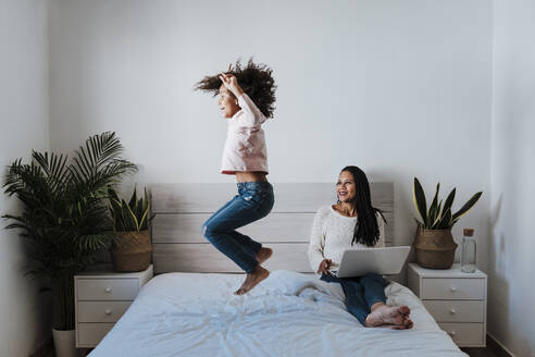 Daughter jumping on bed while mother using laptop at home - EBBF00935