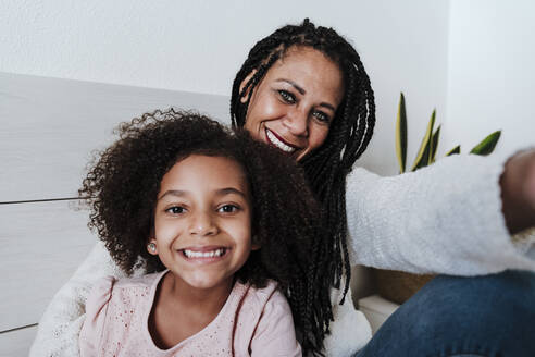 Happy mother and daughter sitting on bed in bedroom at home - EBBF00937