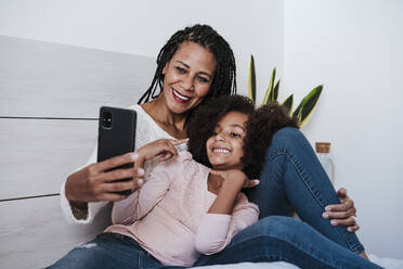 Mother and daughter taking selfie on smart phone sitting in bedroom at home - EBBF00940