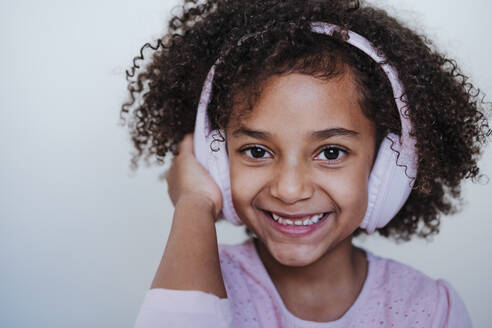 Girl listening music through headphones while standing against wall at home - EBBF00954