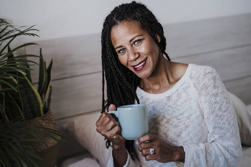 Lächelnde reife Frau trinkt Kaffee, während sie zu Hause sitzt - EBBF00956